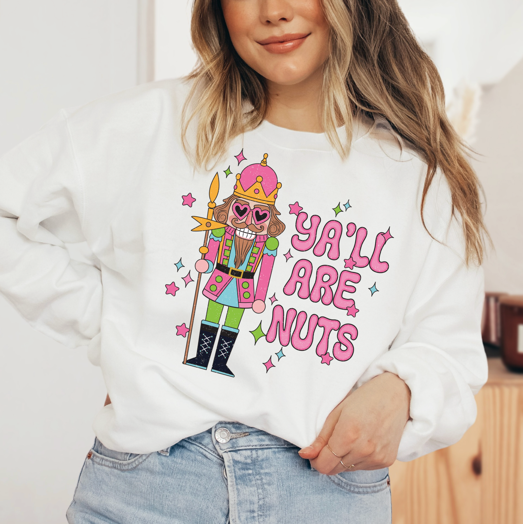 A woman wearing a white sweater with a colorful graphic design of a nutcracker character and the text "Ya'll are nuts" printed on it.