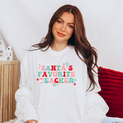 A woman wearing a white t-shirt with the text "Santa's Favorite Teacher" printed on it, smiling at the camera.