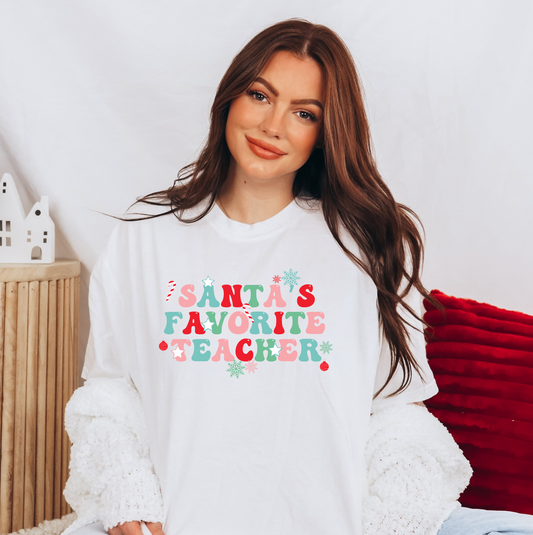 A woman wearing a white t-shirt with the text "Santa's Favorite Teacher" printed on it, smiling at the camera.