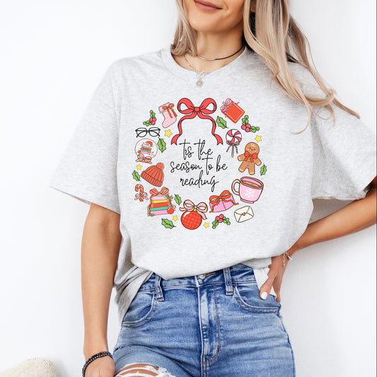 A woman wearing a grey t-shirt with a graphic design featuring various Christmas-themed items such as a wreath, presents, and other festive decorations.