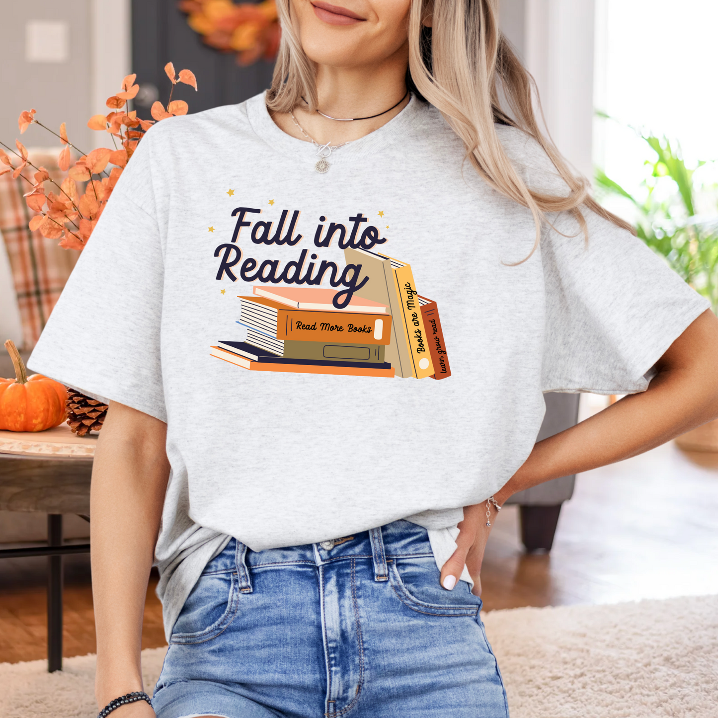 A woman wearing a white t-shirt with a graphic design of stacked books and the text "Fall into Reading" is standing in a room with a pumpkin decoration and a plant.