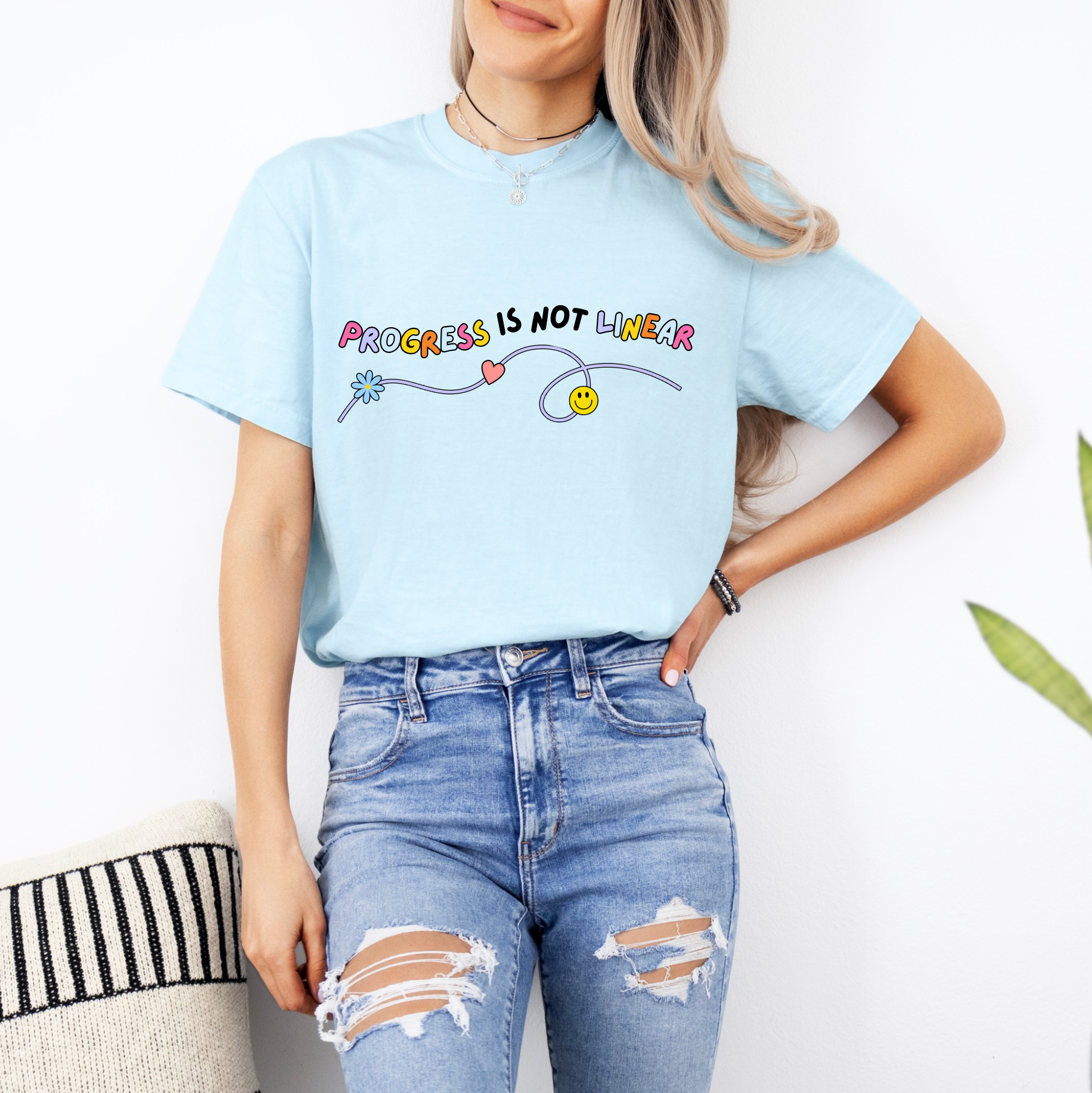 A woman wearing a light blue t-shirt with a colorful graphic and text, paired with distressed blue jeans, stands against a white wall.