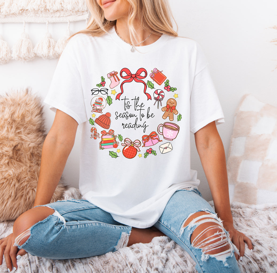 A woman wearing a white t-shirt with a wreath design and text printed on it, sitting on a bed with a beige blanket.