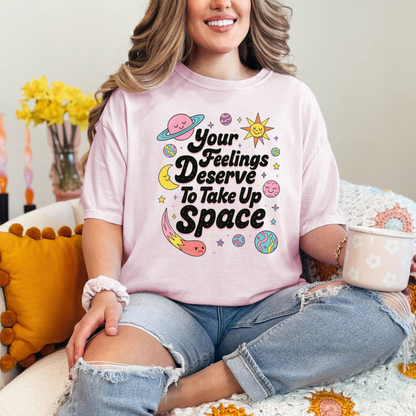 A woman wearing a pink t-shirt with a colorful graphic design and text, sitting on a couch with a cup in her hand.