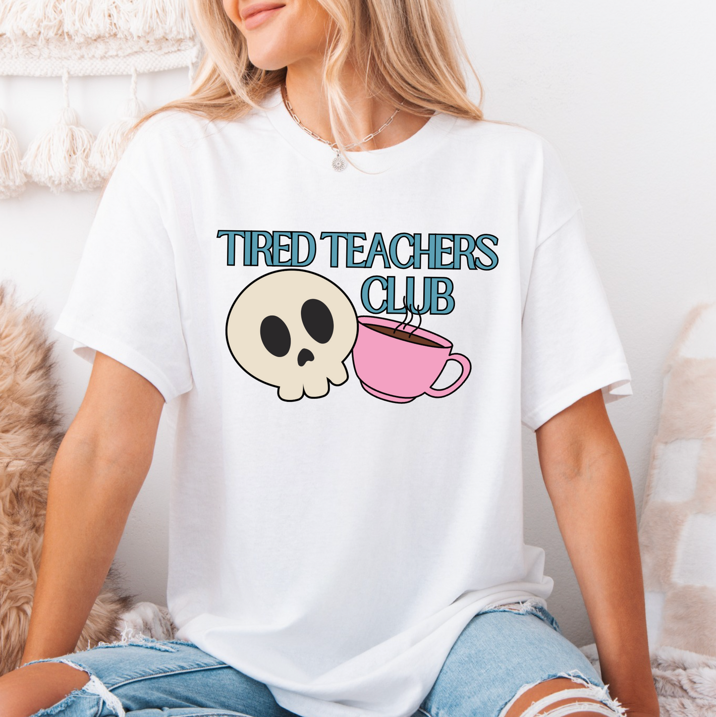 A woman wearing a white t-shirt with a graphic design that says "Tired Teachers Club" and features a skull and a pink coffee mug.