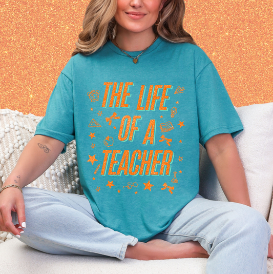 A woman wearing a teal t-shirt with the text "THE LIFE OF A TEACHER" printed on it, sitting on a couch.