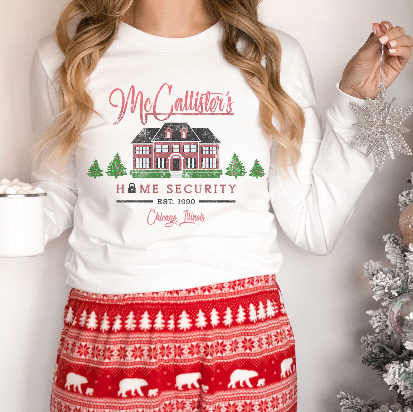 A woman wearing a white long-sleeved shirt with a graphic design of a house and trees, along with the text "McCallister's Home Security Chicago Illinois" printed on it. She is also holding a snowflake ornament.