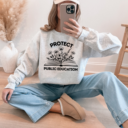 A person is sitting on the floor, taking a selfie with a smartphone, wearing a grey sweatshirt with a graphic design and text.