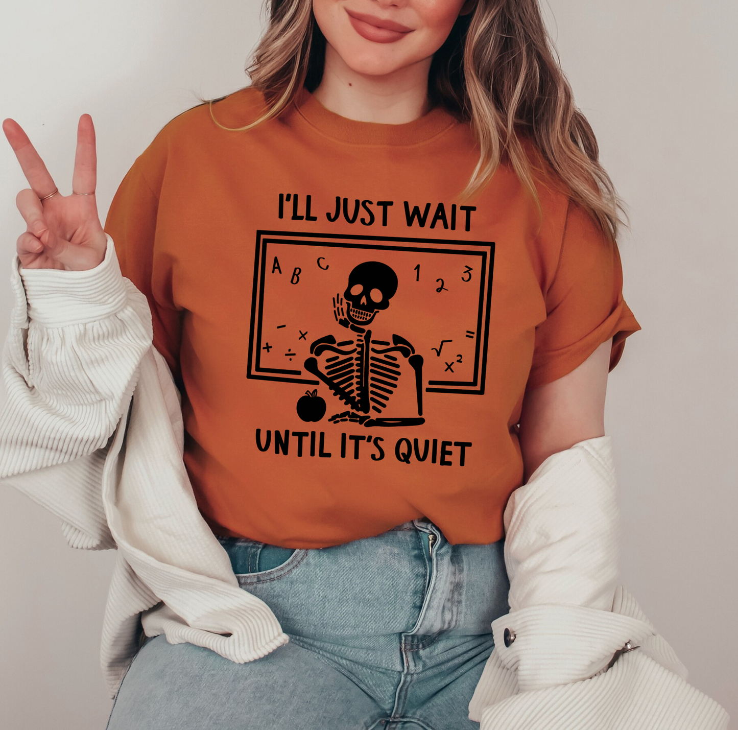 A woman wearing a t-shirt with a skeleton graphic and text, smiling and making a peace sign gesture.