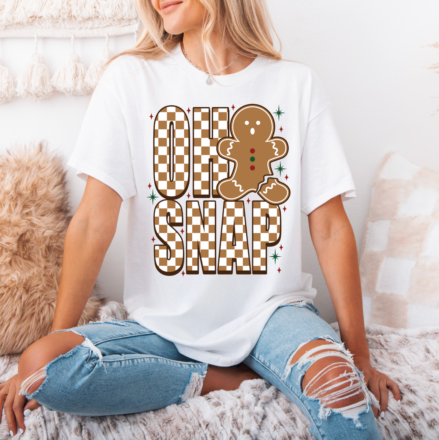 A woman wearing a white t-shirt with a graphic design of a gingerbread man and the text "OH SNAP" is sitting on a bed.