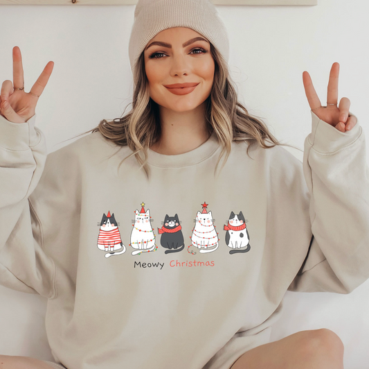 A woman wearing a white beanie and a gray sweatshirt with a graphic of five cats and the text "Meowy Christmas" is making a peace sign with her hands.