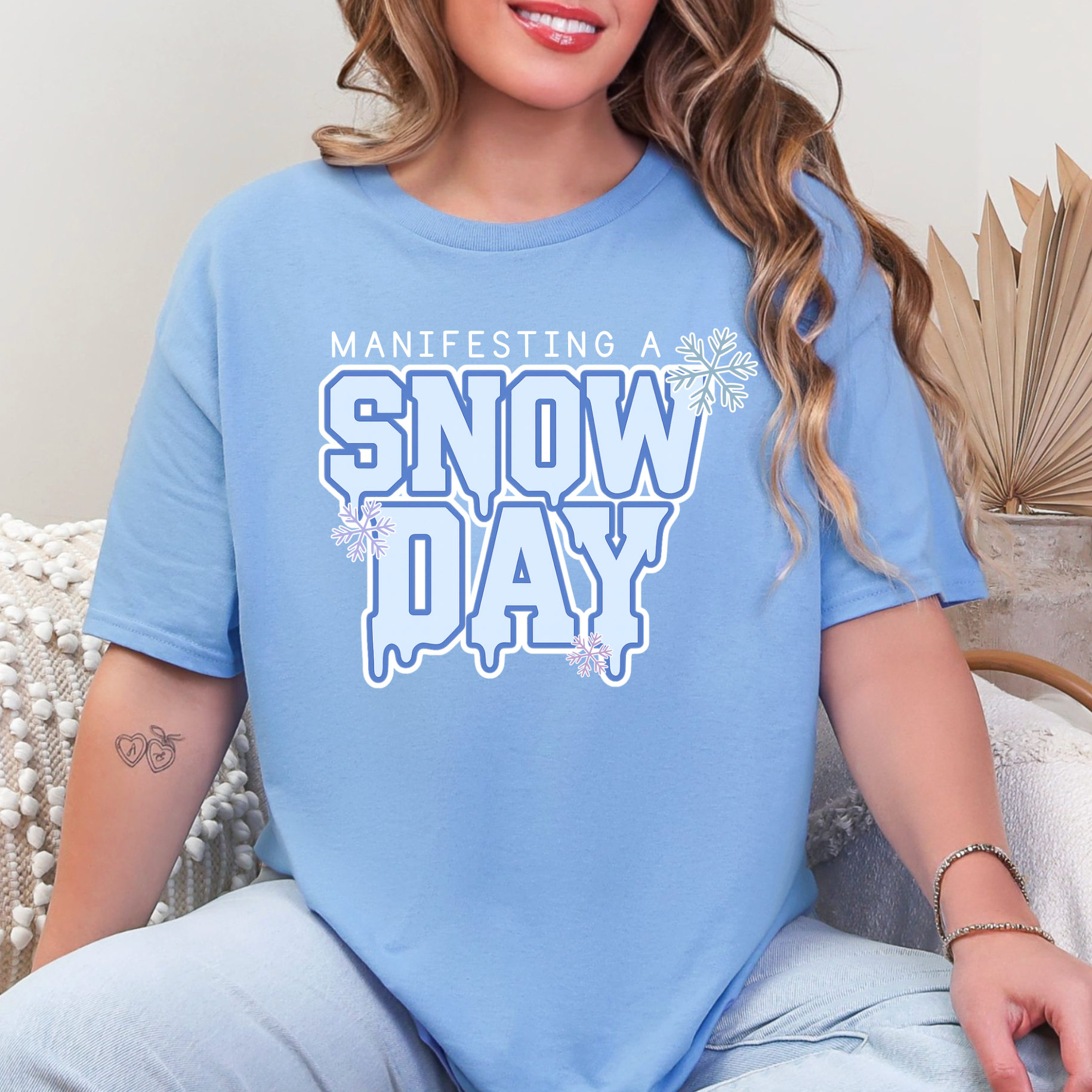 A woman wearing a light blue t-shirt with the text "MANIFESTING A SNOW DAY" printed on it.
