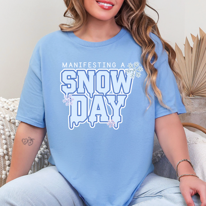 A woman wearing a light blue t-shirt with the text "MANIFESTING A SNOW DAY" printed on it.