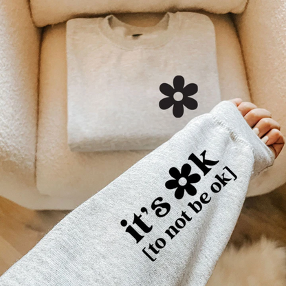 A person's arm wearing a gray sweatshirt with the text "it's ok [to not be ok]" printed on it, along with a black flower graphic.