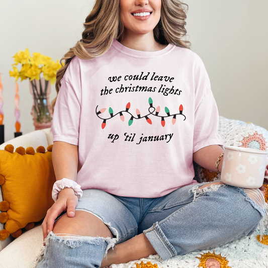 A woman wearing a pink t-shirt with a Christmas-themed design and text, sitting on a couch with a cup in her hand.