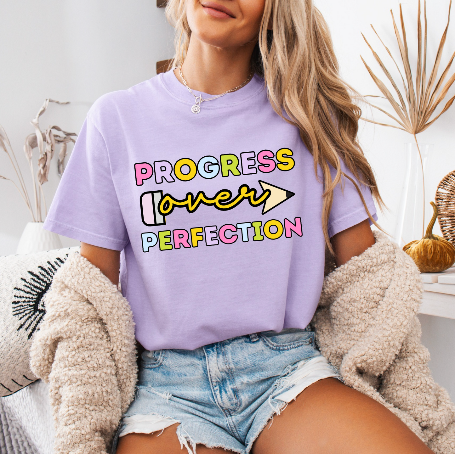 A woman wearing a purple t-shirt with the text "PROGRESS OVER PERFECTION" printed on it, sitting in a room with a beige jacket and a plant in the background.
