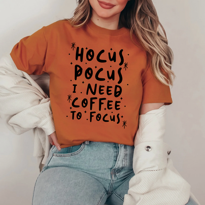 A woman wearing a brown t-shirt with the text "HOCUS POCUS I NEED COFFEE TO FOCUS" printed on it.