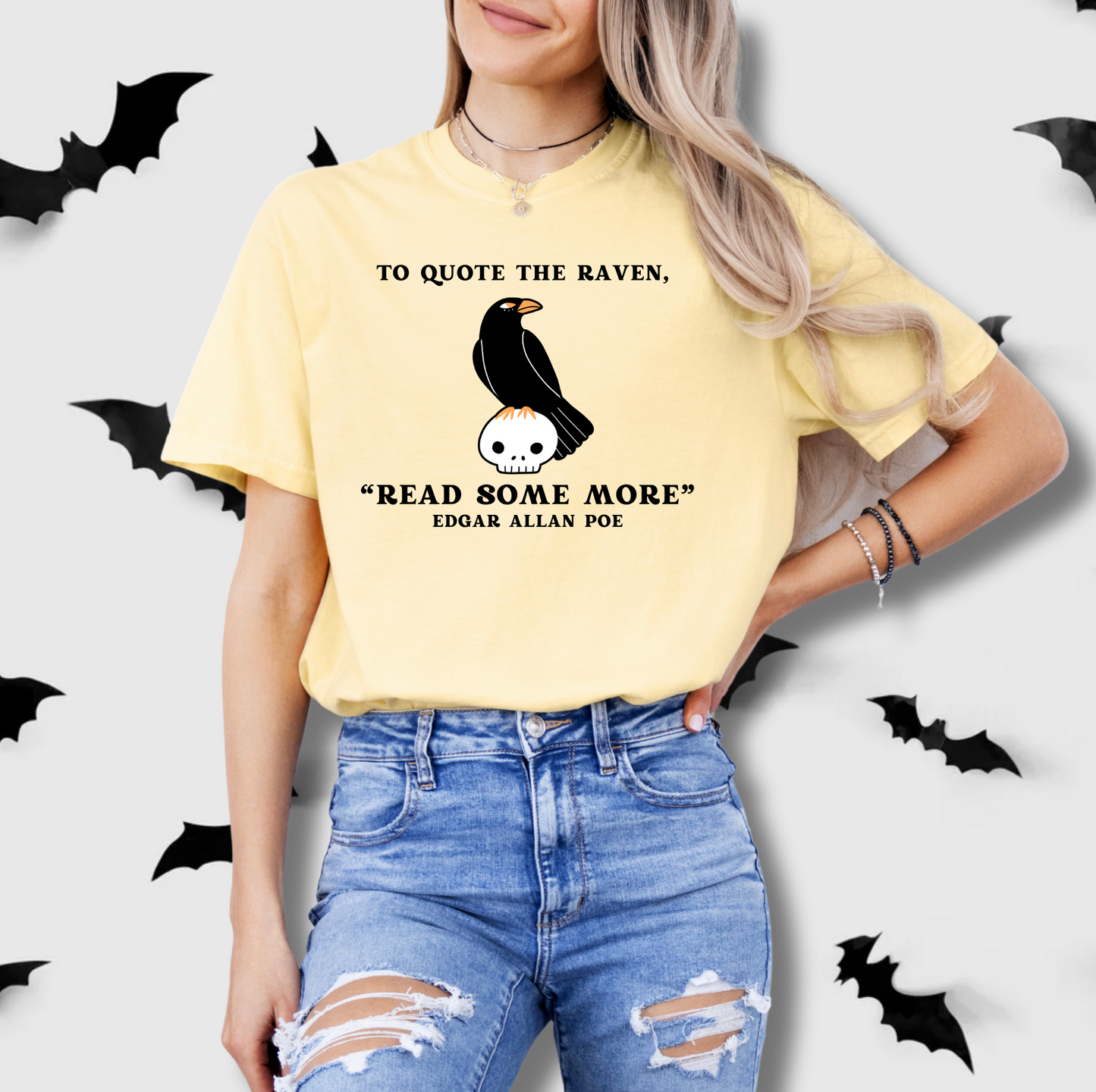 A woman wearing a yellow t-shirt with a quote and a graphic of a black bird on a skull, standing against a background of bats.