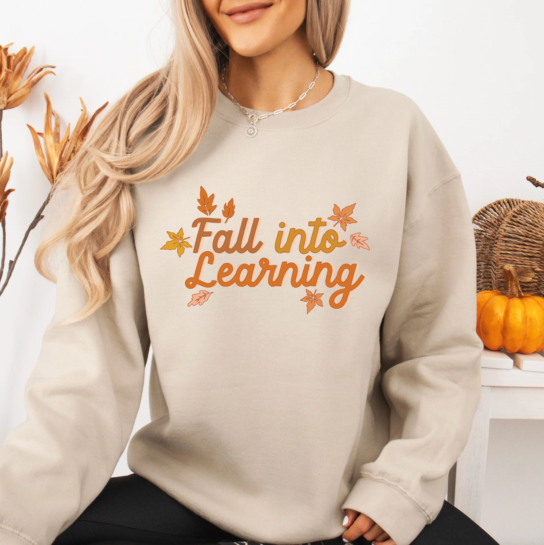 A woman wearing a grey sweatshirt with the text "Fall into Learning" printed on it, sitting in front of a white wall with autumn decorations.