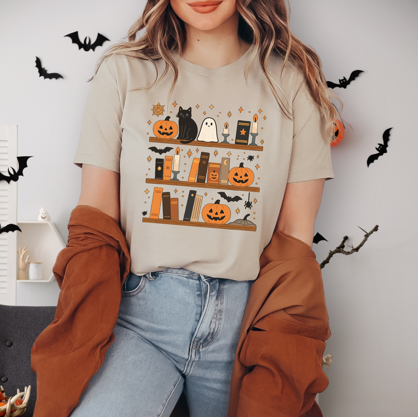 A woman wearing a t-shirt with a Halloween-themed graphic of books and bats, standing in front of a wall decorated with bats and pumpkins.