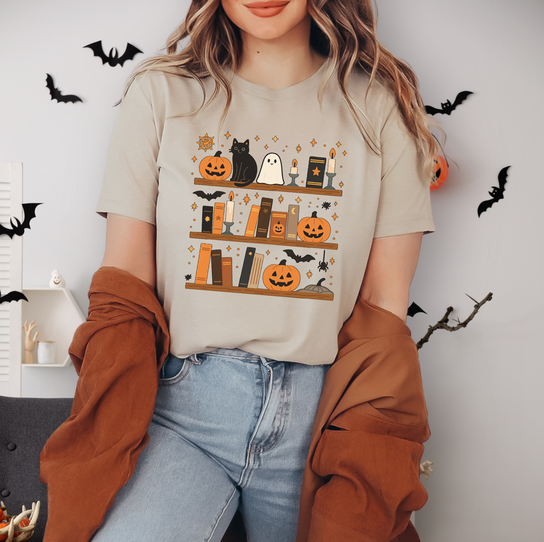A woman wearing a t-shirt with a Halloween-themed graphic of books and bats, standing in front of a wall decorated with bats and pumpkins.